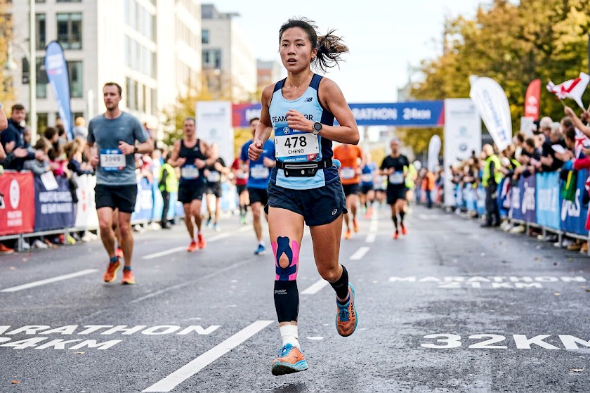 A female athlete is running at full speed on a marathon course; she has pink kinesiology tape applied to her knees and calves and white athletic tape wrapped around her ankle, set against the backdrop of a bustl